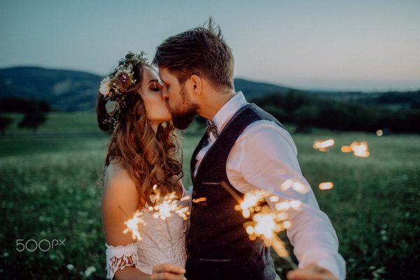 Beautiful young bride and groom on a meadow in the evening, holding sparklers. A man and a woman kissing.