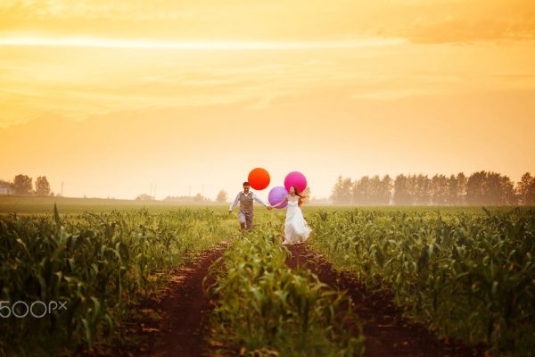 Happy young wedding couple running on the sunset field with big bright colorful balloons